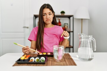 Obraz na płótnie Canvas Young brunette woman eating sushi using chopsticks pointing down looking sad and upset, indicating direction with fingers, unhappy and depressed.
