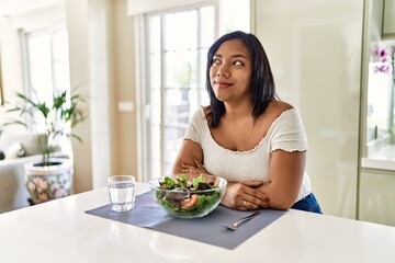 Obraz na płótnie Canvas Young hispanic woman eating healthy salad at home smiling looking to the side and staring away thinking.