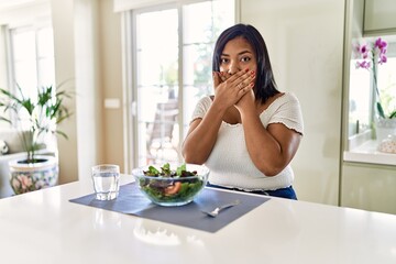 Young hispanic woman eating healthy salad at home shocked covering mouth with hands for mistake....