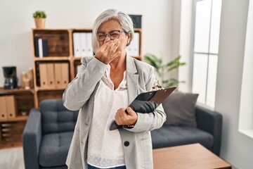 Middle age woman with grey hair at consultation office smelling something stinky and disgusting, intolerable smell, holding breath with fingers on nose. bad smell - obrazy, fototapety, plakaty