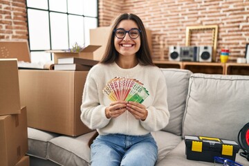 Young hispanic woman sitting on the sofa at new home holding money celebrating crazy and amazed for...