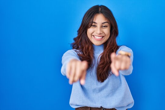 Hispanic young woman standing over blue background pointing to you and the camera with fingers, smiling positive and cheerful
