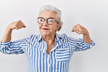 Senior woman with grey hair standing over white background showing arms muscles smiling proud. fitness concept.