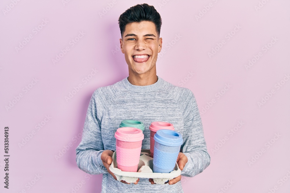 Wall mural young hispanic man holding tray with take away coffee sticking tongue out happy with funny expressio