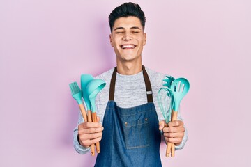 Young hispanic man wearing professional baker apron holding cooking tools smiling and laughing hard out loud because funny crazy joke.
