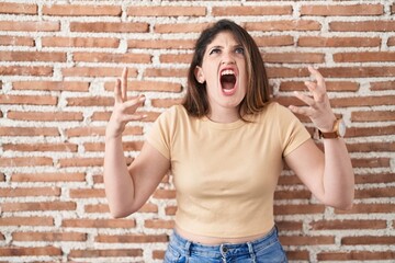 Young brunette woman standing over bricks wall crazy and mad shouting and yelling with aggressive...