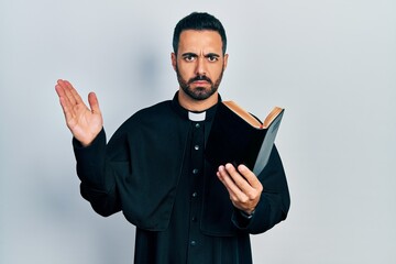 Handsome hispanic priest man with beard holding bible and christian cross skeptic and nervous,...