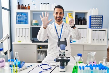 Young hispanic man with beard working at scientist laboratory showing and pointing up with fingers number six while smiling confident and happy.