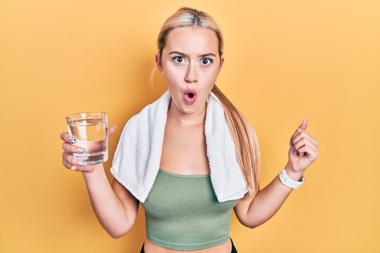 Young blonde girl wearing sportswear drinking glass of water scared and amazed with open mouth for surprise, disbelief face