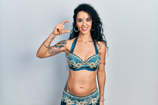 Young woman wearing bindi and traditional belly dance clothes smiling and confident gesturing with hand doing small size sign with fingers looking and the camera. measure concept.