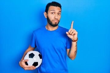Hispanic man with beard holding soccer ball pointing up looking sad and upset, indicating direction...