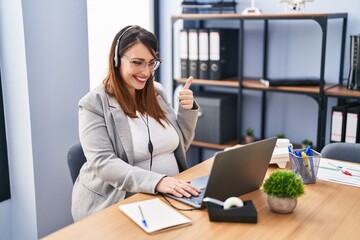 Pregnant woman working at the office wearing operator headset smiling happy and positive, thumb up doing excellent and approval sign