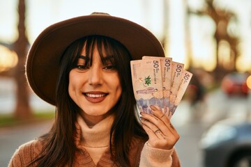 Brunette woman wearing winter hat smiling holding colombian pesos banknotes outdoors at the city on...