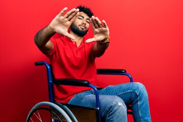 Arab man with beard sitting on wheelchair doing frame using hands palms and fingers, camera perspective