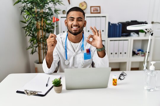Young indian doctor holding reflex hammer doing ok sign with fingers, smiling friendly gesturing excellent symbol