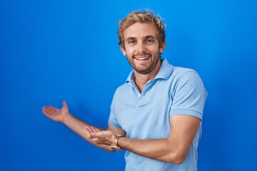 Caucasian man standing over blue background inviting to enter smiling natural with open hand