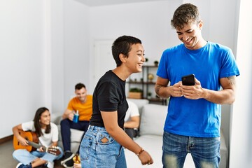 Group of young friends playing classical guitar sitting on the sofa. Man and woman standing and using smartphone at home.