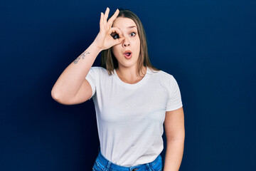 Young hispanic girl wearing casual white t shirt doing ok gesture shocked with surprised face, eye looking through fingers. unbelieving expression.
