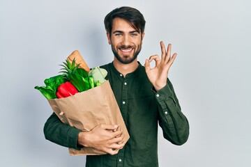 Young hispanic man holding paper bag with bread and groceries doing ok sign with fingers, smiling...