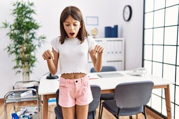 Young hispanic girl standing at pediatrician clinic pointing down with fingers showing advertisement, surprised face and open mouth