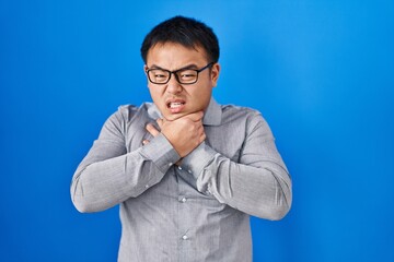 Young chinese man standing over blue background shouting and suffocate because painful strangle. health problem. asphyxiate and suicide concept.
