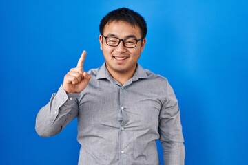 Young chinese man standing over blue background showing and pointing up with finger number one while smiling confident and happy.