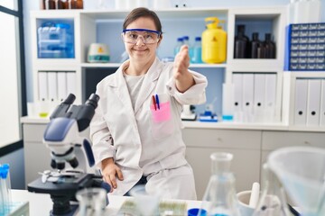 Fototapeta premium Hispanic girl with down syndrome working at scientist laboratory smiling cheerful offering palm hand giving assistance and acceptance.