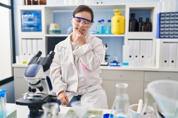 Fototapeta premium Hispanic girl with down syndrome working at scientist laboratory looking confident at the camera smiling with crossed arms and hand raised on chin. thinking positive.