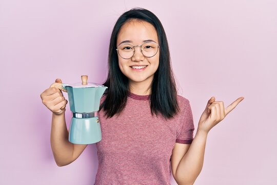 Young chinese girl holding italian coffee maker smiling happy pointing with hand and finger to the side