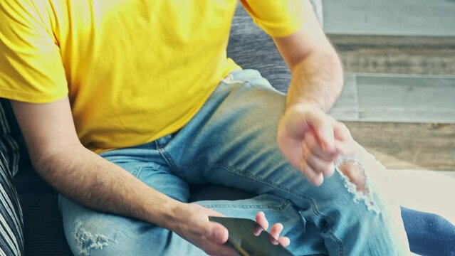 unrecognizable young dark-skinned boy in a yellow t-shirt and blue jeans using his smartphone sitting on a sofa