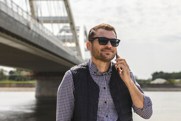 Casual clothing man in sunglasses calling mobile phone and smiling while standing near a big metal bridge above the river.