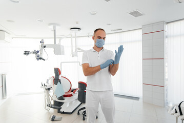 A young dentist in a mask stands near a red dental chair and smiles in modern white dentistry. Modern dentistry and prosthetics.