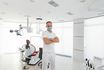 Fototapeta premium A young dentist in a mask stands near a red dental chair and smiles in modern white dentistry. Modern dentistry and prosthetics.