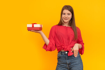 Smiling young girl holds gift box in palm of her hand on bright yellow background. Portrait of woman with gift