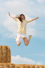 Laughing girl jumps from high haystack. Happy young woman has fun on the farm on blue sky...