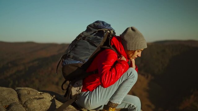 slow motion adventurous woman hiker in red jacket with backpack sits on cliff top at sunset above the woods, gets ready to going and stands up after rest. forward with renewed vigor.