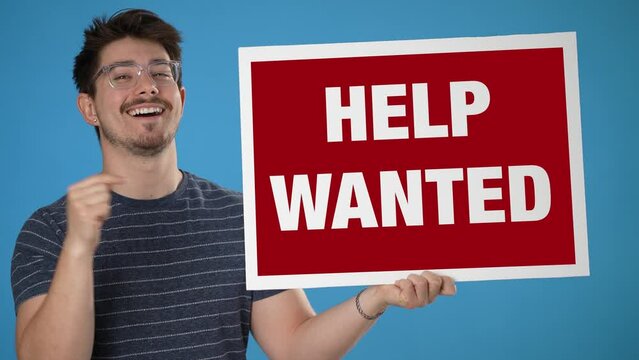 Excited smiling happy trendy man holding large Help Wanted sign on solid blue background.