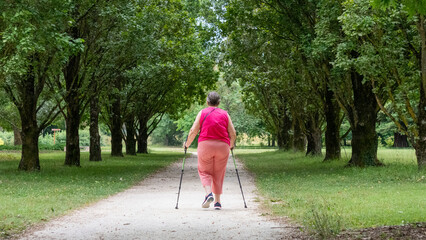 Woman in pink, from behind walking with sticks, on a leafy, green path