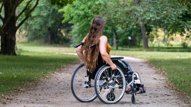 Young woman with long hair, moving in a wheelchair, in profile, in the middle of an alley lined with tall green trees