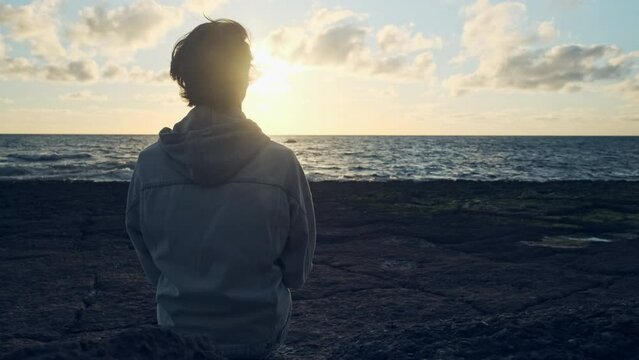 Human looks to the sun over horizon while sunrise. Man on a beach is looking distance during  summer sunset.  Person contemplates the beauty of nature. Freedom. Slow motion. Cinematic dramatic look.