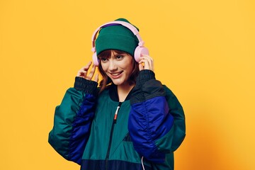 a happy, joyful woman in a green jacket and hat enjoys music standing in pink headphones on a yellow background holding them with her hands and looking away with a pleasant smile on her face