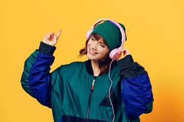 a happy, joyful woman in a green jacket and hat enjoys music standing in pink headphones on a yellow background raising her hand up