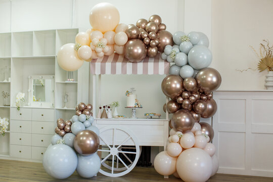 Street vending cart against the backdrop of an indoor cabinet, decorated with brown, beige and gray helium balloons, flowers. On the cart is a cake with the inscription Happy Birthday, ice cream.