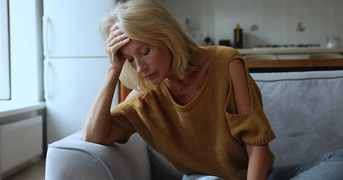 Concerned nervous senior mature woman feeling stress, sitting on couch, holding head, suffering from headache, emotional crisis, depression, anxiety, going through loss, mental disease, disorder