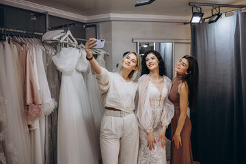 Beautiful bride is trying on an elegant wedding dress in modern wedding salon, her friends are helping