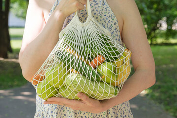 Knitted mesh bag with vegetables and fruits in the hands of a woman. Zero waste grocery shopping concept. - obrazy, fototapety, plakaty