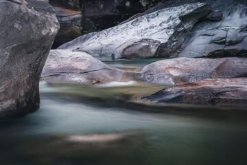 Fototapeta premium Atmosphärischer Naturhintergrund mit riesigen Steinen im Bergfluss. Große Felsen im mächtigen Wasserstrom, Nahaufnahme. Naturhintergrund mit Wald. Dunkel türkisblauer Fluss mit Steinen.