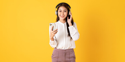 Cute young asian teenage woman using smartphone wearing headphone isolated on yellow background