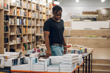 African american man picking and reading books in library or bookstore - obrazy, fototapety, plakaty