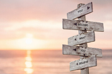 you become what you believe text engraved on wooden signpost by the ocean during sunset. - obrazy, fototapety, plakaty
